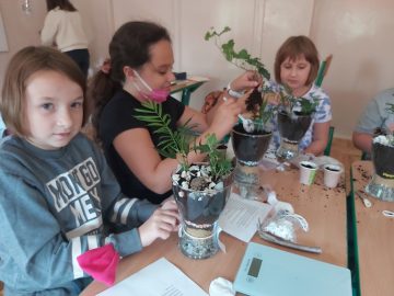 trzy uczennice klasy czwartej siedzące w klasie przy wspólnym stole, tworzące kompozycje kwiatowe w szklanych doniczkach
